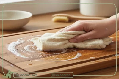دستی در حال تمیز کردن وسایل چوبی آشپزخانه، به ویژه یک تخته برش چوبی، با یک دستمال و آب صابون.